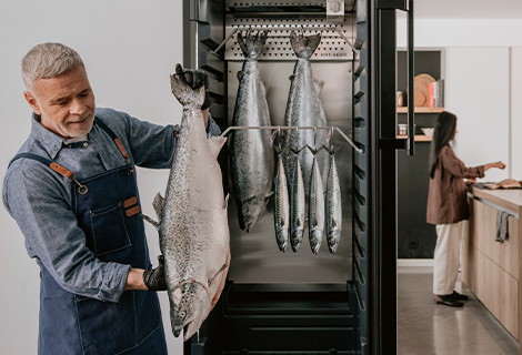 Mann hält einen großen Fisch neben einem DRY AGER Reifeschrank, in dem mehrere Fische zur Reifung hängen.